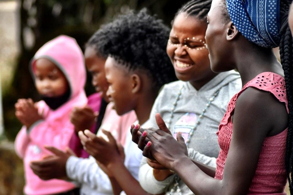 A group of children and young women are smiling and clapping together.