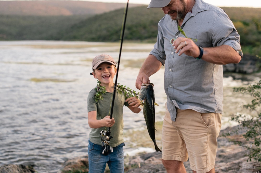 A boy and a man are fishing together by a river, with the boy smiling and holding a fishing rod and a fish.
