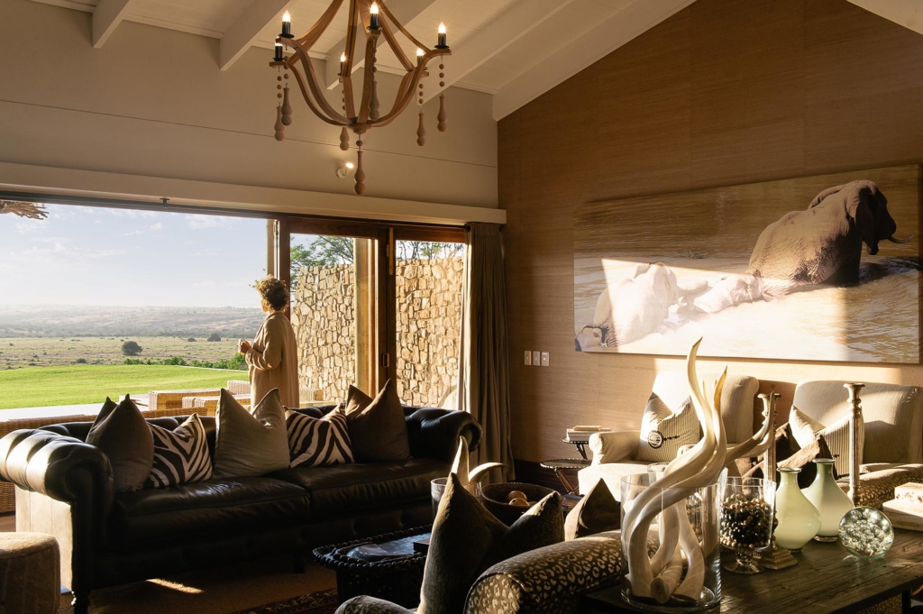A woman stands near an open window in a stylish living room with elegant furnishings and a view of the landscape.