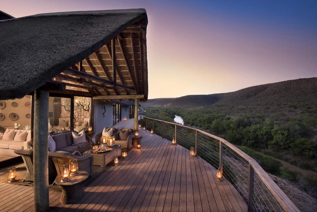 A wooden deck with stylish furniture and lanterns overlooking a river and hills at sunset.
