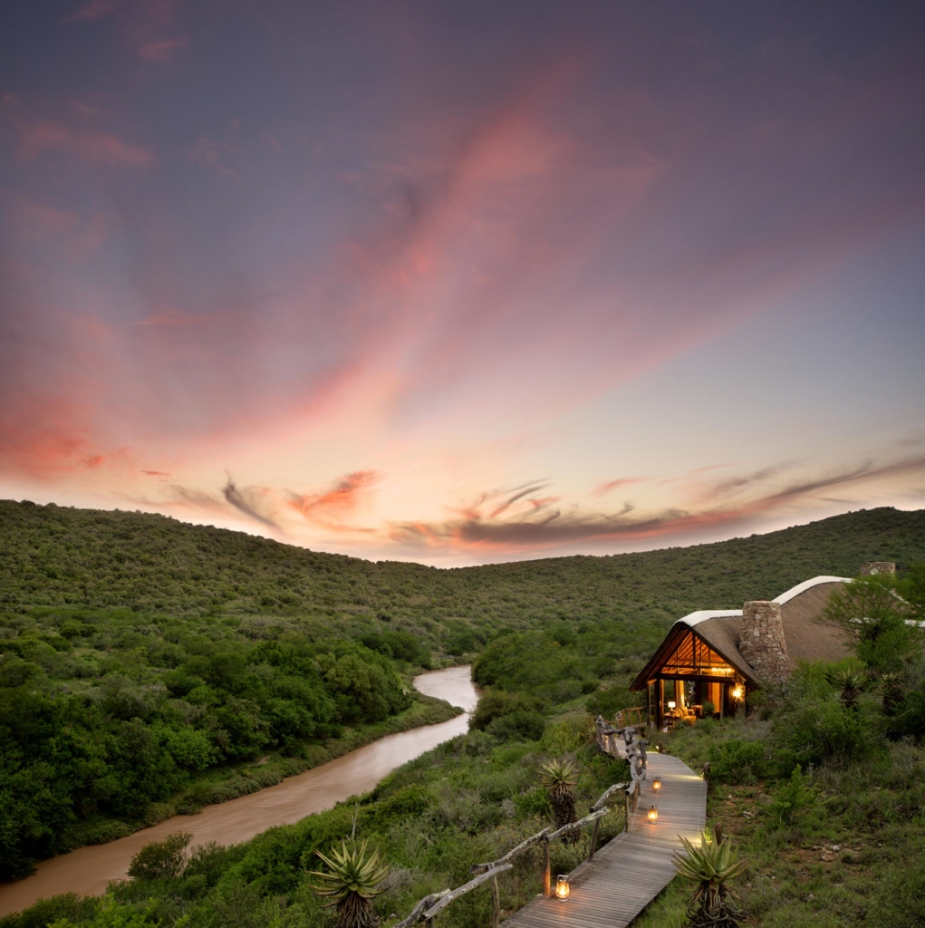 A lodge sits beside a winding river with hills in the background under a colorful sky at sunset.