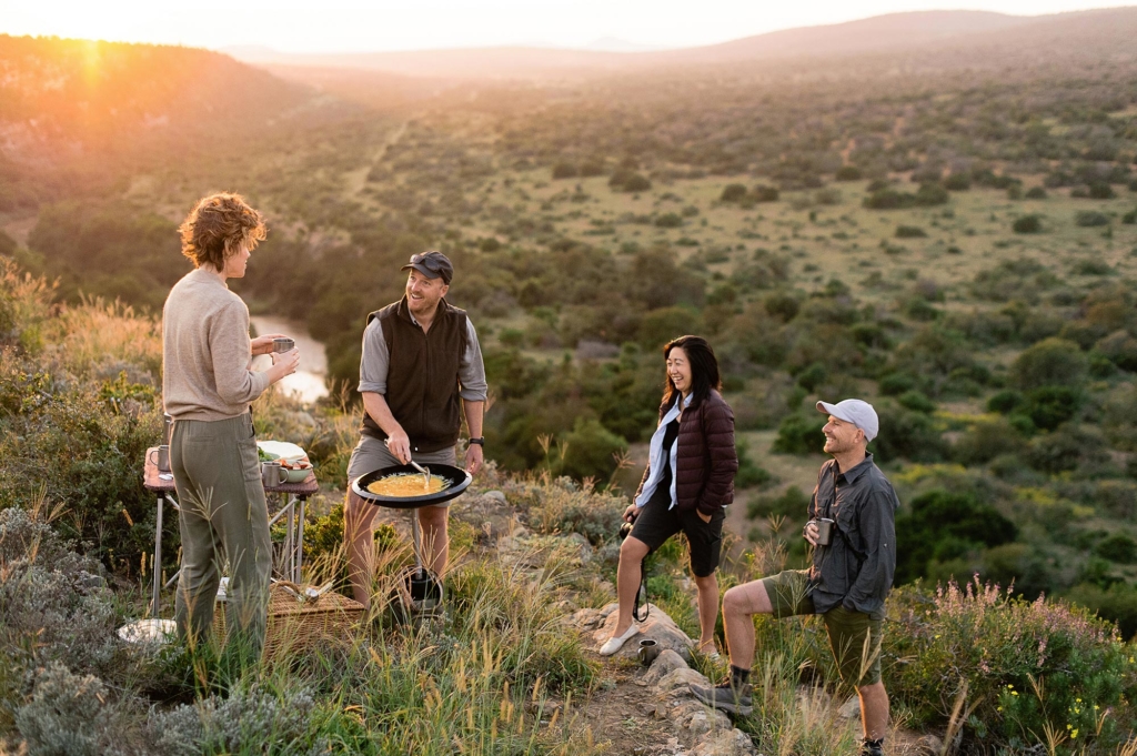 A group of four people is enjoying a barbecue on a hillside during sunset.
