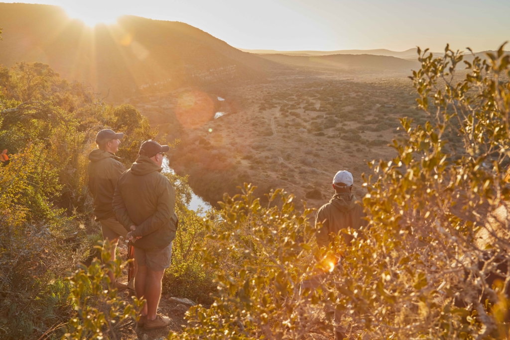 Three people standing on a hillside overlooking a valley at sunrise.
