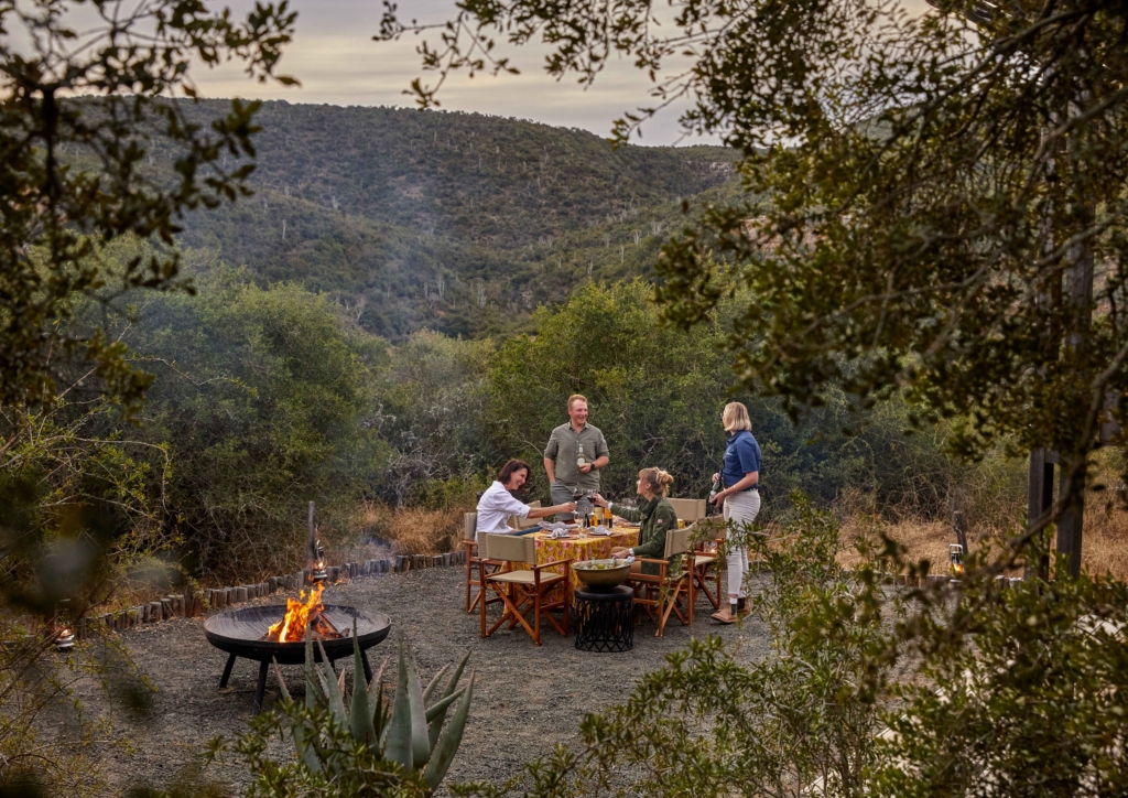 A group of people enjoy a meal around a fire in a scenic outdoor setting.