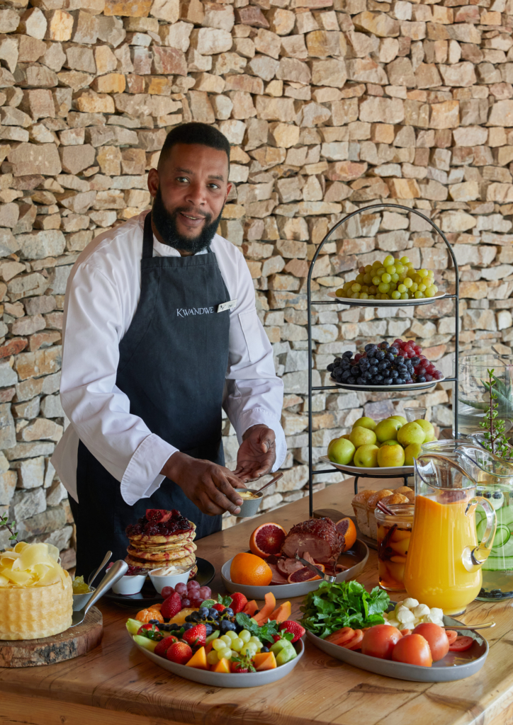 A chef stands at a wooden table filled with a variety of fresh fruits and dishes.