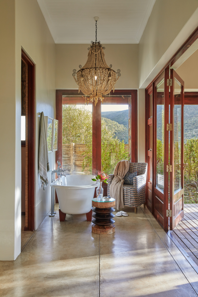 A luxurious bathroom with a chandelier, a freestanding tub, and views of nature through large windows.