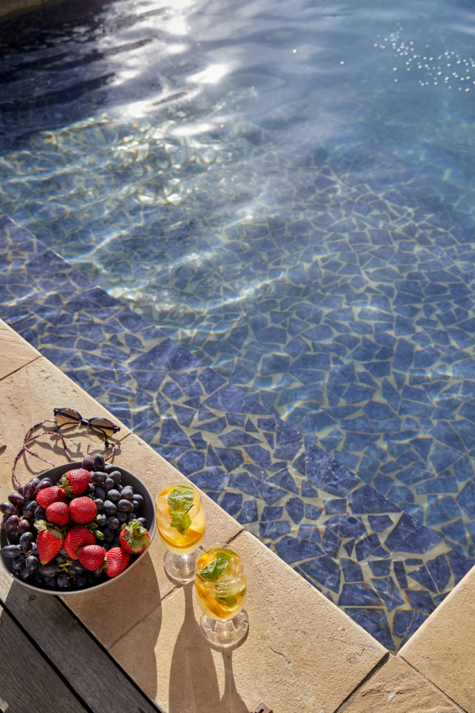 A bowl of mixed berries and two cocktails sit beside a shimmering blue pool.