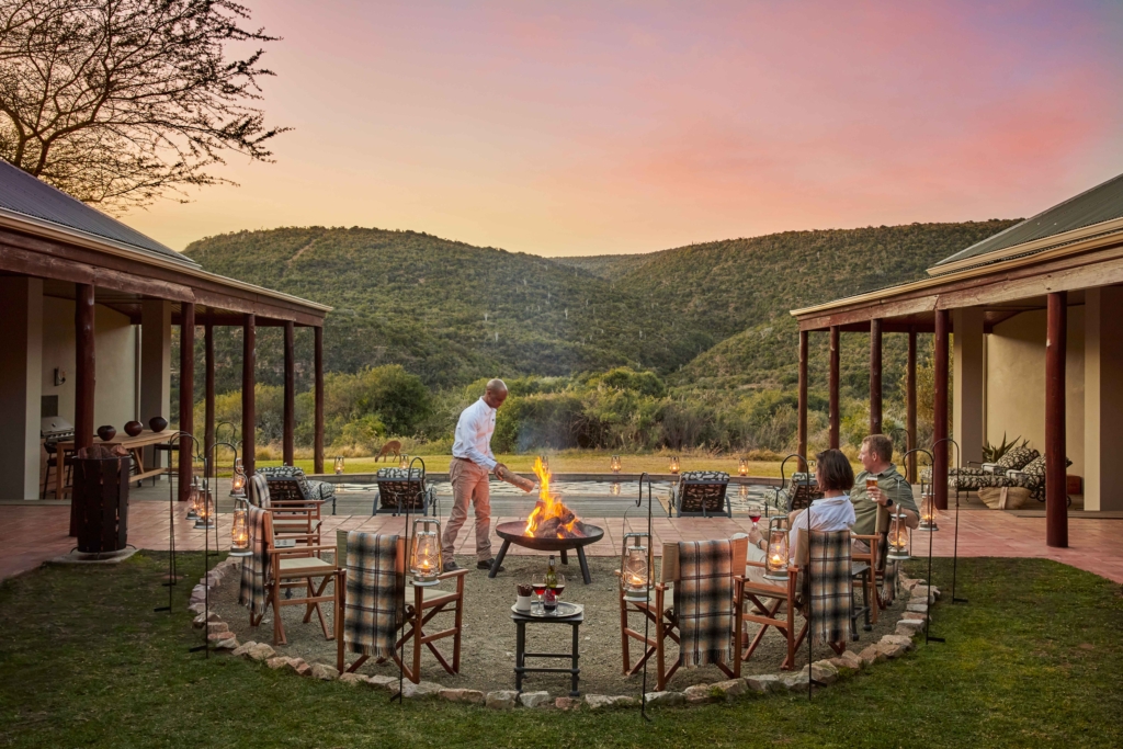 A group of people gathers around a fire pit in a scenic outdoor setting at sunset.