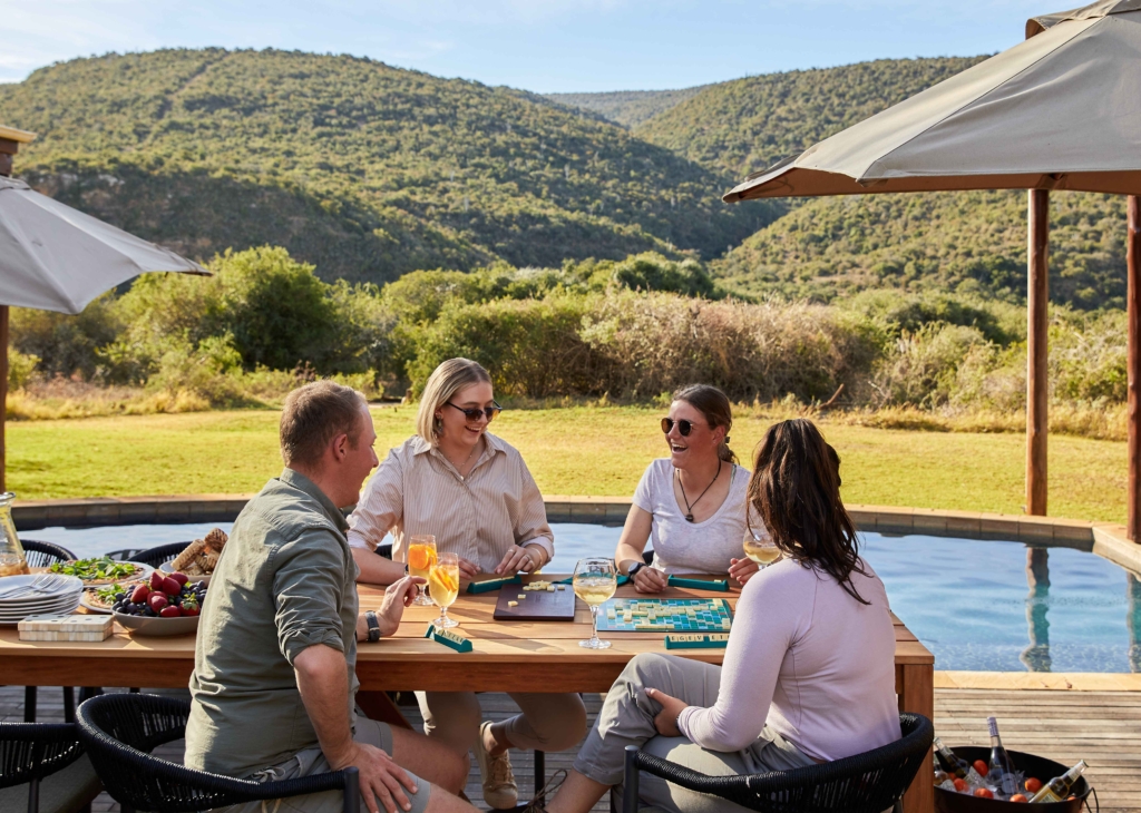A group of four people enjoying drinks and games at an outdoor table near a pool with mountains in the background.