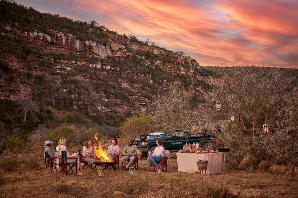 A group of people gathered around a campfire in a scenic outdoor setting during sunset.