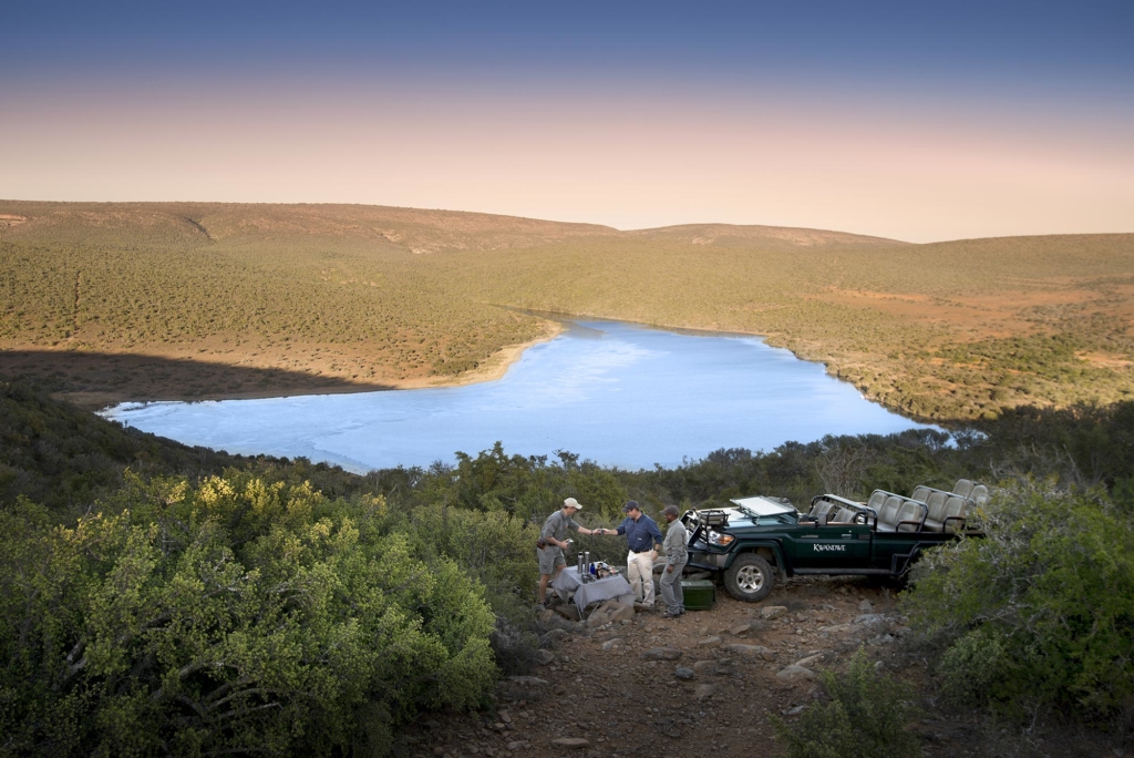 A group of people near a vehicle by a lake surrounded by greenery and hills.