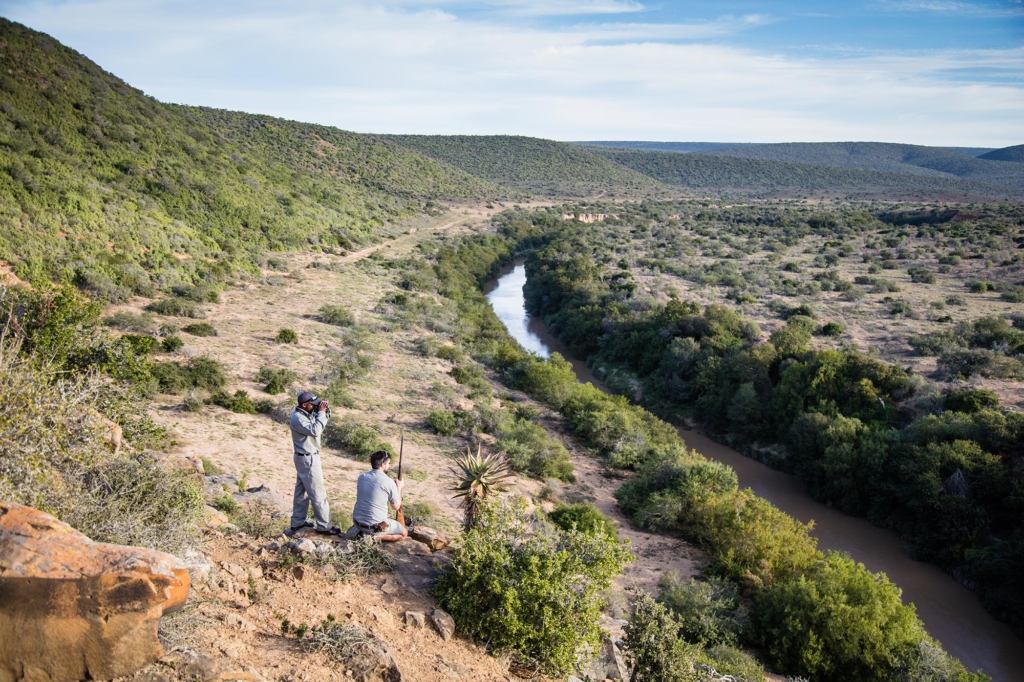 Two people observe a winding river from a hillside surrounded by greenery.