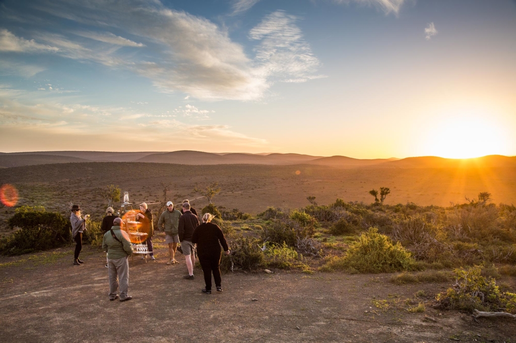 A group of people stands on a hillside overlooking a landscape at sunset.