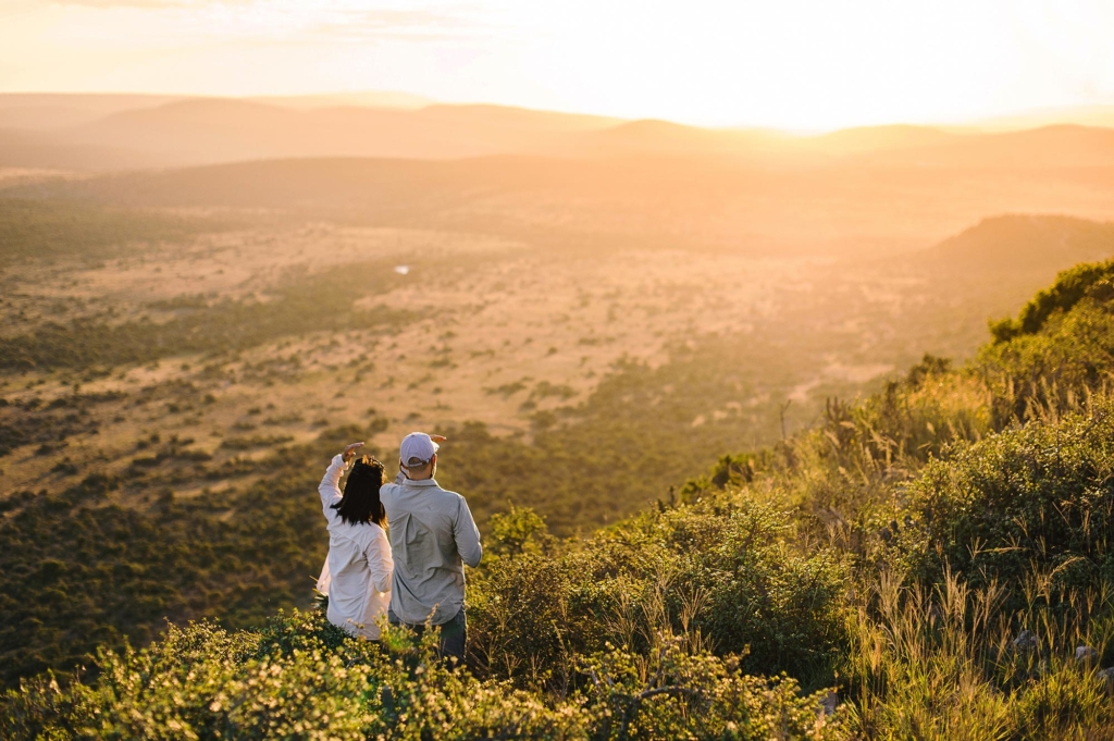 A couple stands on a hilltop overlooking a vast landscape at sunset.