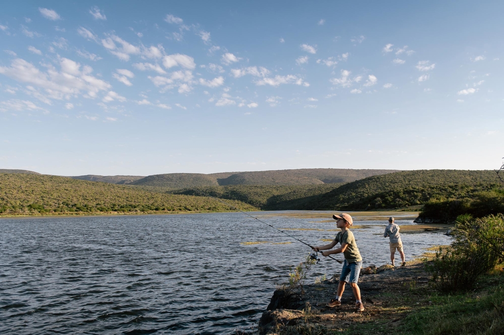 A child and an adult are fishing by a lake surrounded by green hills under a partly cloudy sky.