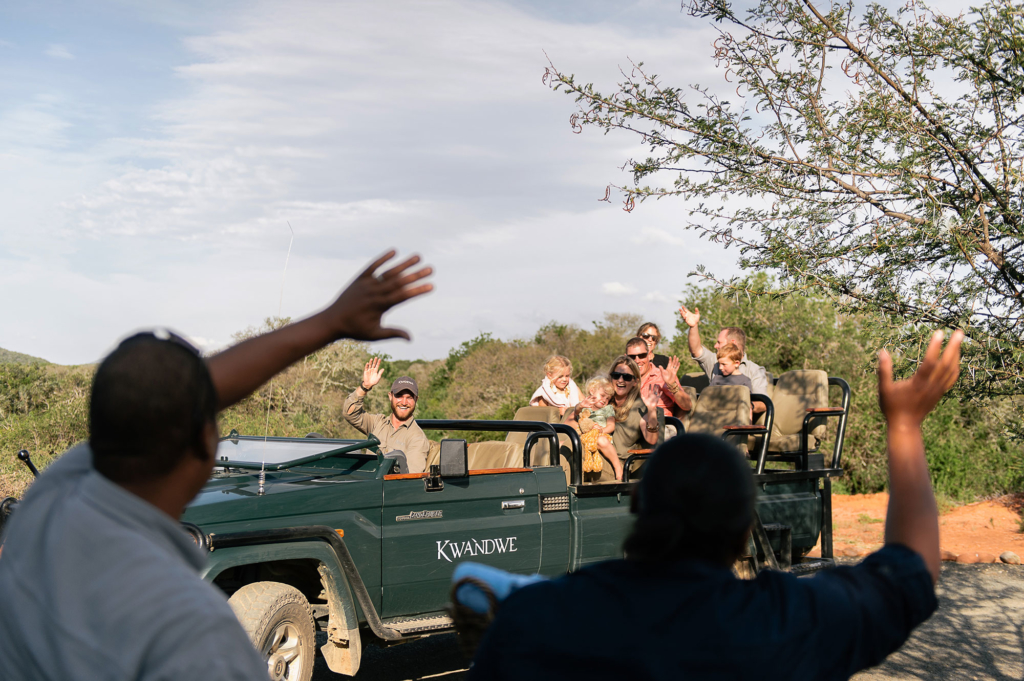 People waving from a safari vehicle in a natural setting.
