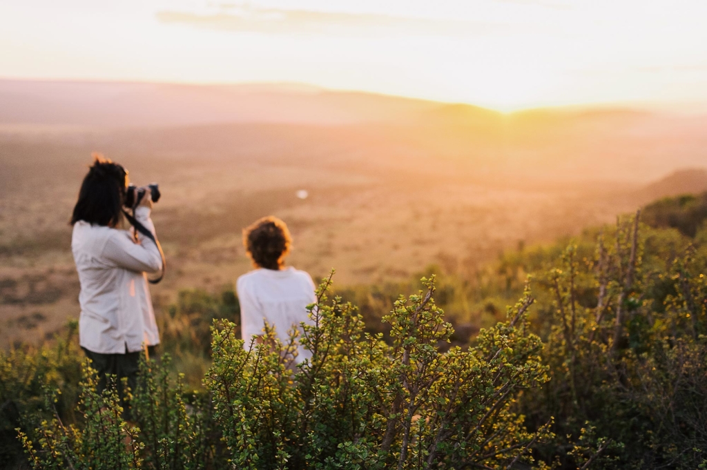 Two people are observing a sunset while one photographs the landscape.