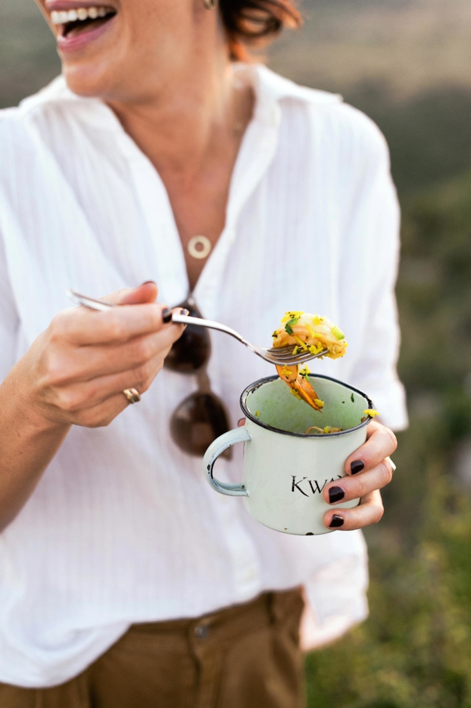 A person is smiling while holding a fork and a cup with food in an outdoor setting.