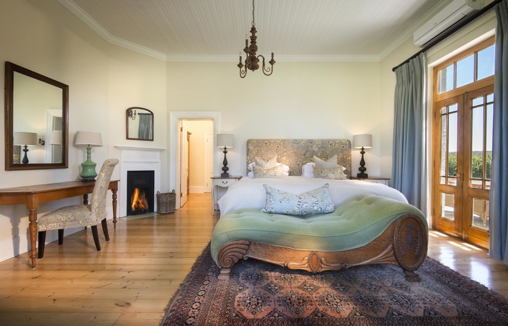 A spacious, elegantly furnished bedroom featuring a large bed, a desk, and windows with natural light.