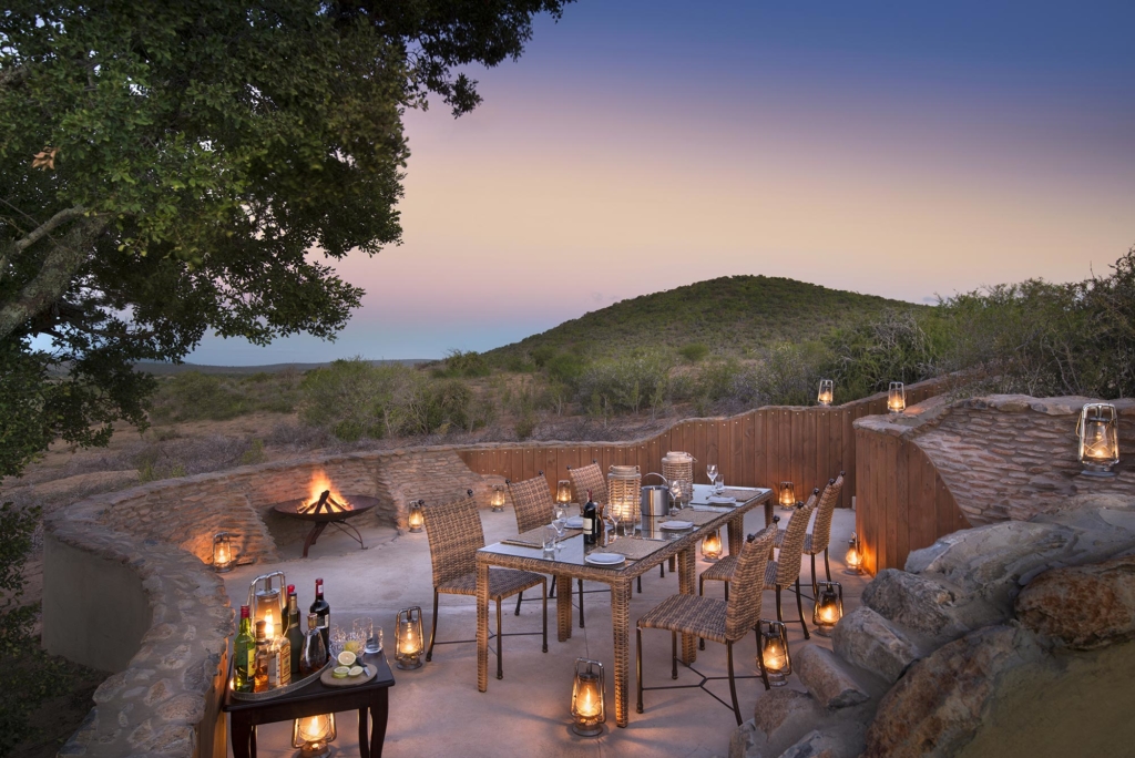 An outdoor dining area is set up on a terrace with lanterns and a fire pit, surrounded by hills at dusk.