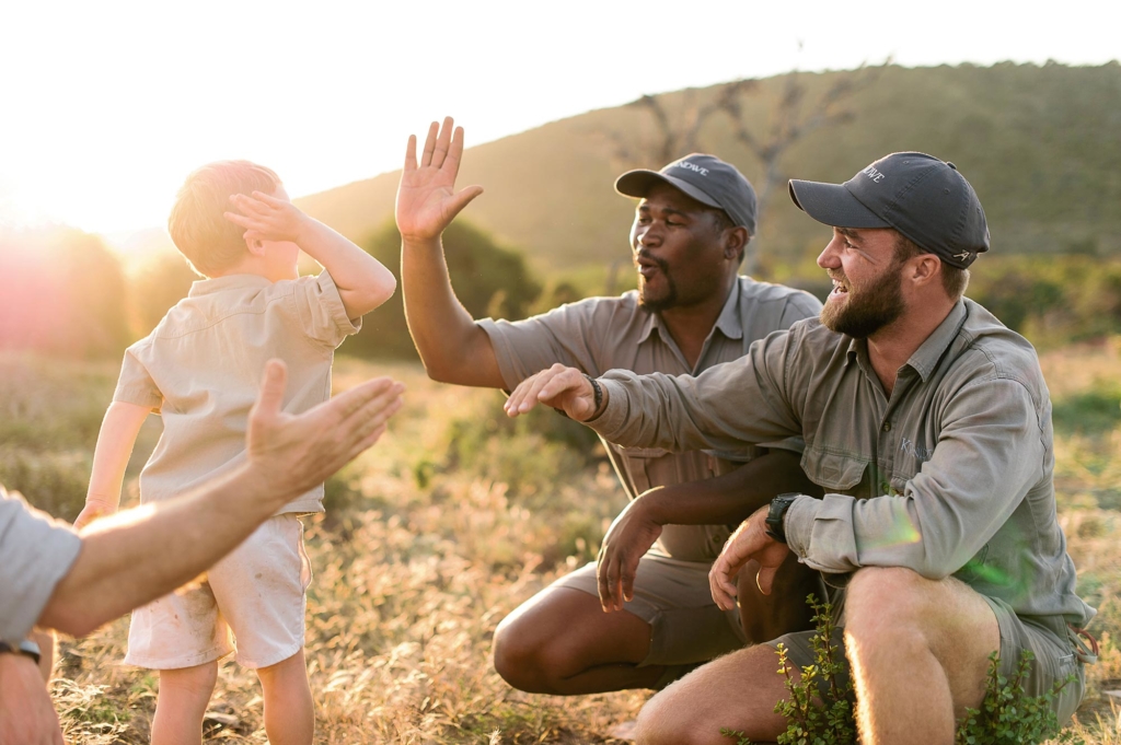 Two adults greet a child in a natural outdoor setting during sunset.